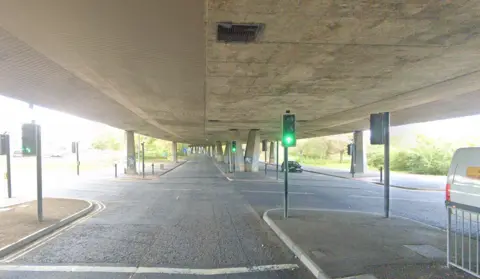 Google A huge concrete flyover held up by many pillars sits above two roads. There are traffic lights on green and a car is approaching them.