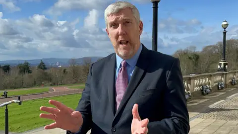 PA Media A man wearing a dark suit stood in front of parliament buildings in Belfast