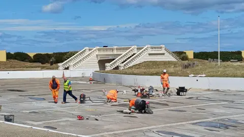 Redcar Model Boat Club Five men in high visibility orange clothing and white helmets are repairing the concrete floor of the lake, surrounded by building equipment and materials. There is a white ornamental footbridge at the back of the lake. The sky is blue. 