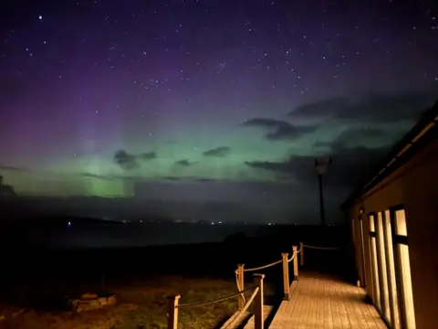 Craig Nisbet The Northern Lights from a deck. A house is on the right with some lights on. 