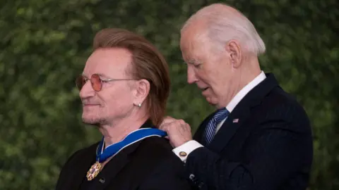 Bloomberg/Getty Images Side profiles of singer Bono wearing and President Joe Biden. Bono is wearing red-tinted round glasses and a black top. Biden is wearing a dark suit over a white shirt with a navy and white striped tie. He is putting a medal around Bono's neck.