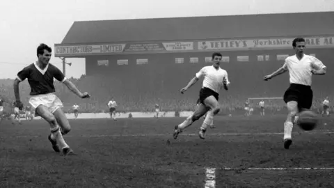 PA Media Dougan watches his shot fly towards the goal in a match in front of a packed crowd. Two rival players wearing white run towards the ball. The pitch is very muddy.