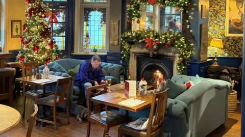 The pub is furnished with a mixture of pale green sofas and wooden tables and chairs. The fireplace is covered in a illuminated garland and a Christmas tree sits in the background. A man sits on a sofa with a stemmed glass of beer in front of him and a dog lying at his feet in front of an open fire.