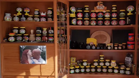 Marmite jars and memorabilia on shelves in a house, with a photo of David and Laura Walker also on the side