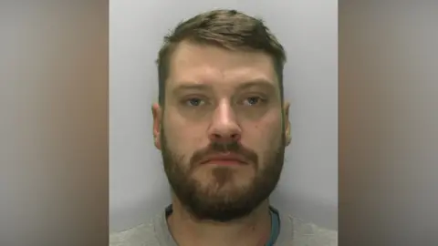 Custody image of Jordan Ireland. He is wearing a grey sweatshirt and has brown hair and a beard.