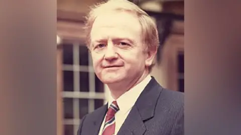 Kevin Hickey with ginger hair, white shirt, red, white and black club tie and dark grey  suit