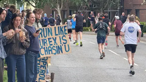 Spectators watching the race from the side of the road. One person holds a sign which says 'RUN LIKE UR EX IS CHASIN' U'