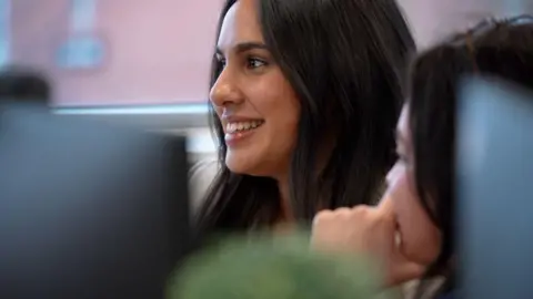 Faith Khan is looking at her computer screen whilst smiling. She has long dark hair. The foreground is blurred but another woman can just be made out from behind. She has her hand to her face while watching Khan.