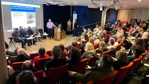 Martin Giles/BBC People sit on auditorium seats and face the stage, where four people are sitting down, with one person in a purple shirt talking, with a slideshow presented behind him.