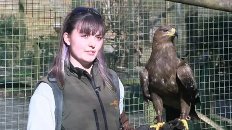 Bird team member Jennie Marshall with Stilton the steppe eagle. They are in front of his enclosure. He is resting on her left arm.