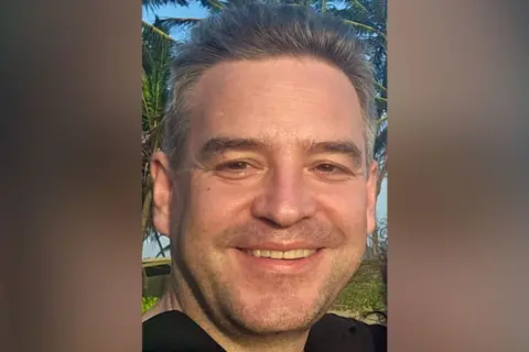 John Ames closeup of face, he has greyish hair, in the background are tropical trees.