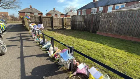 BBC A path in front of terraced houses which is lined with flowers, balloons and tributes.