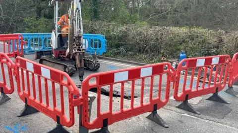 A worker wearing orange sits to operate a small roadworks vehicle, the arm of which is poking into a hole in the road. The area has been surrounded by blue and orange plastic barriers.