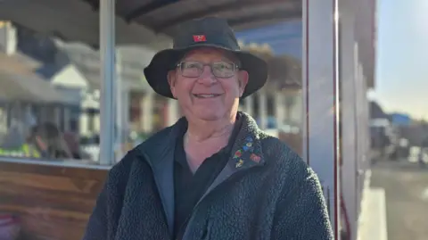 Harry Colville is wearing a dark fleece jacket and a dark bucket hat with a small Manx flag badge on the front. He's an older gentleman and is smiling and wearing glasses, while sitting at the front of a tram carriage on a sunny day.