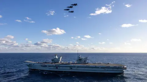 Royal Navy HMS Prince of Wales travels through the sea, with four F-35s flying above it.