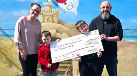 Family The two young boys from the above photo holding a large cheque for £1,000 made out to "Birmingham Children's Hospital Charity". On either side of them are a woman and a man.