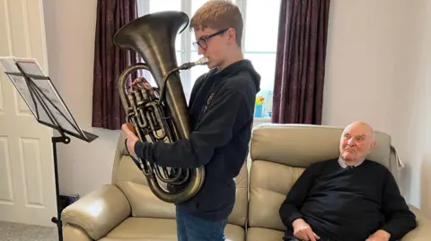 Neythen, a 13-year-old boy, is reading music from a stand and playing a brass euphonium. Behind him, Brian Routledge is sitting on a sofa watching him.