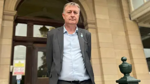 Man with grey hair in grey suit with light blue shirt stood outside a beige stone building with wooden door entrance