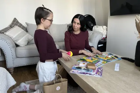 A 10 year old girl with short dark hair, a burgandy top and white joggers kneels at a pale wood coffee table which is covered with felt pens, magazines and story books. A large brown paper gift bag is on the floor next to her. A woman with long dark wavy hair and a burgandy top is the kneeling at the end of the table looking at the child. There is a pale grey sofa behind them.