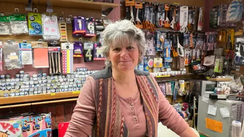 Karen Dawkins is standing behind the counter in her shop. Behind her are miscellaneous items on display.