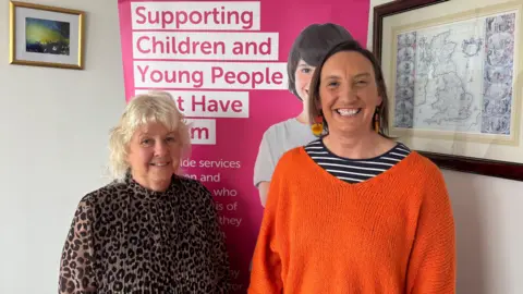 Two ladies smiling at the camera, the one on the left is wearing a leopard print blouse and has white hair and the one on the right has brown short hair and is wearing a black and white shirt which is under a bright orange sweater.