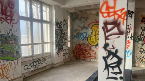 A landscape still of an empty, concrete room. On the right is a large, multi-panel window and there are a number of colourful graffiti tags on the walls and pillars around the room.