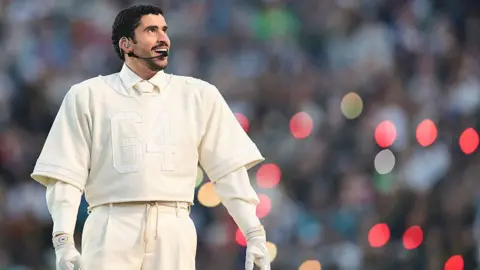 A man dressed all in white - trousers, shirt and tie, gloves, and with a cropped American Football shirt on top. He has a microphone attached to his face and there is an out-of-focus crowd in the background