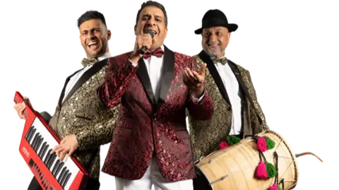 The band are wearing decorative suits, Aamir has a keyboard, Amjad has a microphone and Surinder has a drum. 