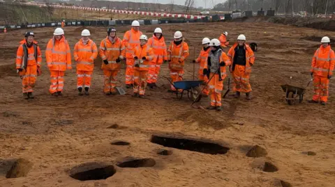 Oxford Cotswold Archaeology A row of 12 archaeologists, dressed in orange hi-vis clothing and white or black hard hats lined up on sandy soil. In front of them are at least 12 excavated Beaker People era pits.