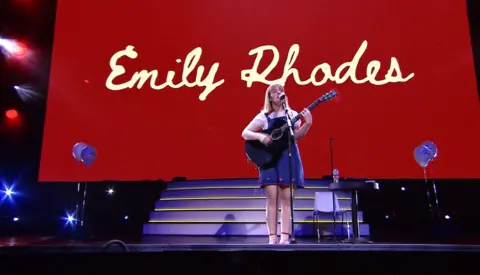 Emily Rhodes Emily Rhodes stands on stage holding a black acoustic guitar and singing into a microphone. On the stage are purple balloons and a purple set of stairs. A large screen is in the background displaying Emily Rhodes written in joined-up font against a dark red background.