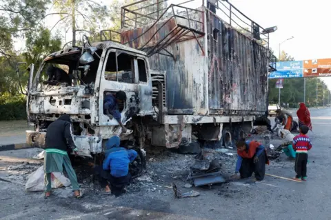 Reuters Women and children collect recyclables from a burnt-out truck used by Bushra Bibi, the wife of jailed former Pakistani Prime Minister Imran Khan. The truck is in the middle of a quiet highway