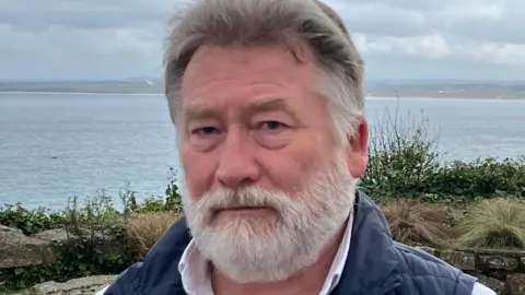 A man with a grey beard in a white shirt and dark blue gilet with the sea in the background 