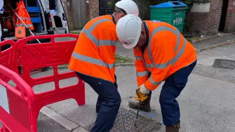 Two workmen have removed a square-shaped manhole cover.