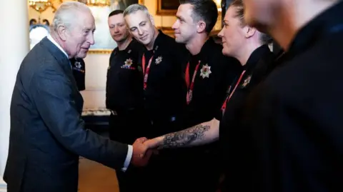 PA Media the king wearing suit, has his hand outstretched shaking the hand of an emergency service worker.