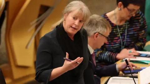 Shona Robison, with blonde hair and wearing a black dress, stands to speak in the Scottish Parliament with a pen in her hand 