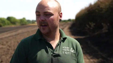 A man with a shaved head is standing beside a cultivated field on agricultural farmland. A ploughed field stretches into the distance, with a hedge along the right side. The man is wearing a dark green polo shirt with a logo which reads NETHER HAUGH FARM.