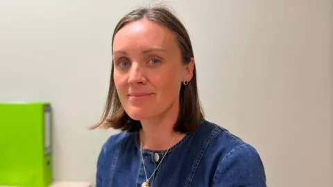 A woman with brown shoulder length hair sitting in an office, wearing a blue denim top with silver buttons. She has on a silver necklace and silver and blue stud earrings. There is a cream wall behind her and a lime green folder on the left of the photo on her desk.