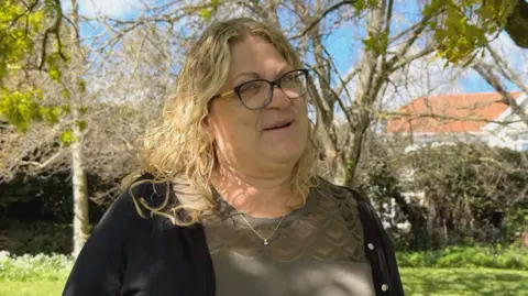 A woman with curly blonde hair and glasses stands in a sunlit garden or park, wearing a green top and black cardigan, with trees and grass around her