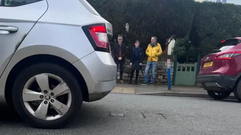 Three campaigners stand on a pavement the opposite side of the road to the camera. A silver car is driving off to the left, while a maroon car is driving off the the right. The campaigners include a man on the left and right with a 12 year-old girls standing between them. 