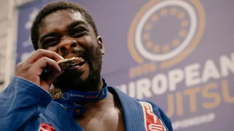 A man biting his medal. He is wearing a blue robe.