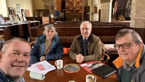 Bridget Weaving Four people sit at a table in a church, with pews and an organ in the background. They have sheets of paper, notebooks and mugs on a table. Three are men, and one a woman. The man taking the picture is smiling and has short fair hair. The collar of his checked shirt can just be seen. The woman has grey bobbed hair with a fringe and is wearing a blue puffer coat with beige jumper. To her right is a man white white hair and glasses wearing a beige jacket and tie. To his right is a man with dark hair and black-framed glasses in a coat with orange collar, green v-neck jumper and blue checked shirt.
