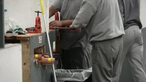 Three anonymous male prisoners wearing grey clothing operate trade tools.