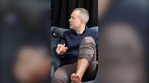 Dr Chris van Tulleken is looking to his right. He is wearing brown trousers. His left trouser leg is pulled up to show his foot.