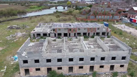 A drone view of two large, half-built apartment buildings standing on overgrown green land beside a wide river. The two-storey shells are open to the elements, with no door, windows or proper roofs. In the background stands a large Edwardian red-brick school with boarded-up windows. 