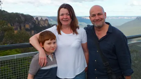 Nicky Wake A family of three stand and pose for a photo on an elevated platform overlooking a scenic hilltop view. A woman with brown hair, Nicky, has her arm around a young boy, with ginger hair, Finn. On Nicky's other side stands Andy with his arm behind hers.