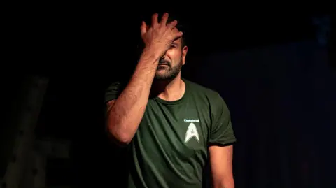Sid Akbar/Bradford Arts Centre A man holds his hand to his face. He is wearing a green T shirt and standing against a black background