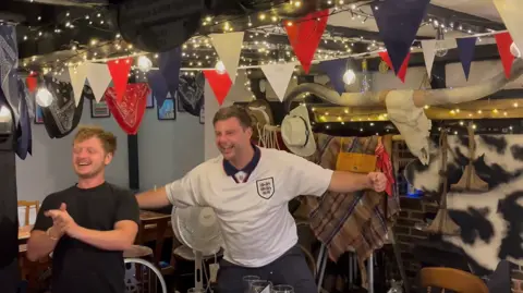 BBC / Phil Harrison Fans in a decorated pub react after an England goal, with two people at the centre of the room clapping and celebrating beneath red, white and blue bunting and string lights.