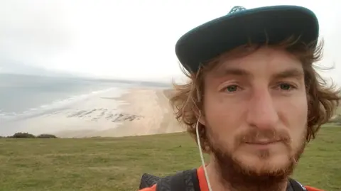 The picture shows a wide coastal view taken from a grassy headland or clifftop. In the foreground, part of a person is visible wearing a dark cap, an orange top and a backpack, with earphones in. In the background, a long sandy beach curves along the shoreline, with the sea stretching out to the left under a pale, cloudy sky. 