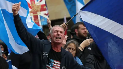 A man raises his fist while standing in front of a group of people waving flags, including saltires and a union flag.