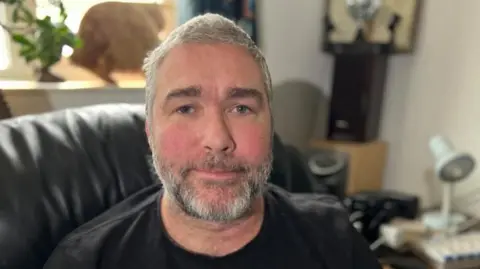 Steve Hubbard/BBC John Goude looks at the camera and wears a black T-shirt. He sits on a leather couch in his living room and has gray hair and a beard